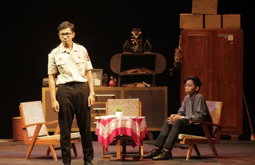 Whatsapp image 20260715 at 17 44 45 catrawarta Man in a police like uniform stands center stage addressing audience two chairs and a pink tablecloth set in a theater scene